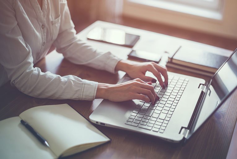 Woman in white shirt typing on a laptop at a wooden desk