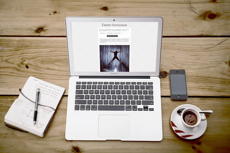 Overhead view of MacBook Air with Embed Instagram page open beside notebook, phone and coffee cup
