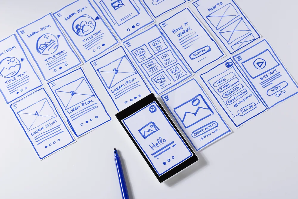 Hand-drawn UI wireframes spread on a desk with a smartphone showing a mockup.
