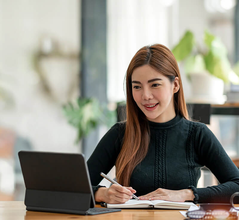 Smiling woman with long brown hair writing in a notebook beside a tablet at a desk